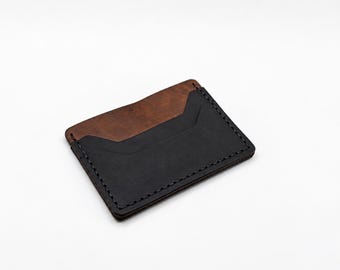 May include: A rectangular leather card holder featuring a two-tone design with a dark brown upper section and a black lower section. The accessory is constructed with visible black stitching along the edges and includes a central slot.