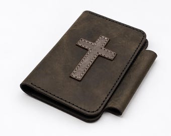 May include: A dark brown leather cover featuring a stitched cross emblem on the front panel. The rectangular cover has visible edge stitching and a side flap, designed to hold a small book or notebook securely in place.