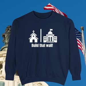 May include: Navy blue sweatshirt with a white graphic of a church, a wall, and a building with a flag on top. The text "Build that wall!" is printed below the graphic.