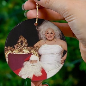 May include: A round ceramic ornament featuring a drag queen in a white dress sitting on a gold throne with a Santa Claus in a red suit sitting on her lap.