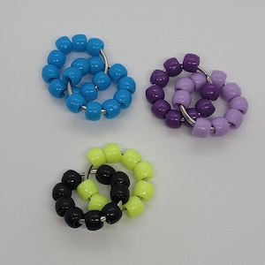 Pode incluir: Três anéis de contas em tons de azul, roxo e preto e amarelo. Cada anel é feito de pequenas contas redondas enfiadas em um anel de metal prateado. Os anéis estão dispostos em uma superfície branca.