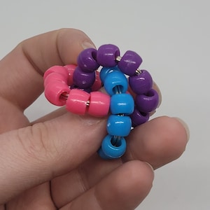 Pode incluir: Um brinquedo de contas fidget colorido com contas roxas, cor-de-rosa e azuis enfiadas em um fio. As contas estão dispostas em espiral.