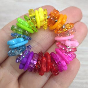 May include: A rainbow-colored bracelet composed of small, translucent plastic rings. The bracelet features a spectrum of colors, including red, orange, yellow, green, blue, and purple. The bracelet is held in a hand.