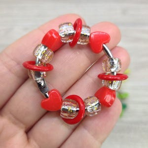 Könnte beinhalten: Ein handgefertigtes Armband mit roten herzförmigen Perlen, transparenten Perlen mit goldenem Glitzer und roten kreisförmigen Perlen. Das Armband ist auf einem silberfarbenen Metalldraht aufgezogen und schafft ein charmantes Accessoire.