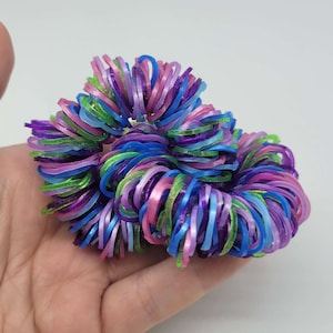 Pode incluir: Um grupo colorido de elásticos em tons de rosa, azul, verde e roxo. Os elásticos estão dispostos em um padrão circular, criando uma forma de flor.