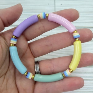 Könnte beinhalten: Ein pastellfarbenes Armband mit vier unterschiedlich farbigen Perlen. Die Perlen sind helllila, hellblau, hellgrün und gelb. Jede Perle hat einen goldenen Akzent und einen dünnen Streifen in einer anderen Farbe.