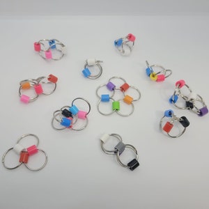 May include: A collection of silver-toned keychains with colorful, geometric beads. The beads are in various colors, including pink, blue, orange, green, and black. The keychains are arranged on a white surface.