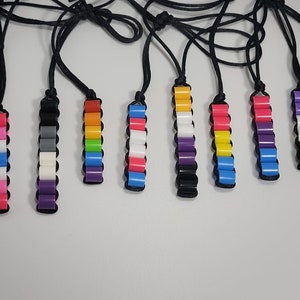 May include: A set of ten colorful beaded necklaces, each representing a different pride flag. The necklaces are made with small, round beads and are strung on black cords.