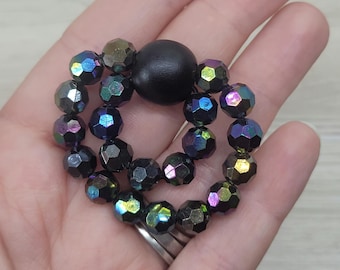May include: A circular beaded accessory featuring iridescent, faceted beads in shades of black, purple, and green. A large, round, black bead sits in the center. The accessory is held in a hand.