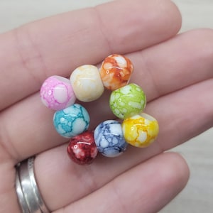 Pode incluir: Uma coleção de nove contas coloridas e marmorizadas em tons de rosa, amarelo, laranja, verde, azul, vermelho e branco. As contas redondas têm um acabamento brilhante. As contas são pequenas, perfeitas para a criação de joias.