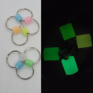 Pode incluir: Dois chaveiros de metal com contas translúcidas coloridas. Um chaveiro tem contas rosa, azuis, laranjas e amarelas. O outro tem contas azuis, verdes e brancas. Uma segunda imagem mostra os mesmos objetos brilhando em verde no escuro.