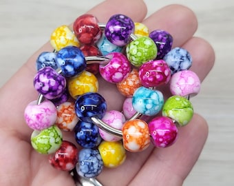 May include: A collection of colorful, marbled beads in various shades of red, purple, blue, yellow, green, pink, and orange, strung on a silver ring. The beads are round and have a glossy finish, creating a vibrant and eye-catching design.