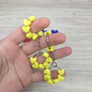 Pode incluir: Dois chaveiros amarelos em forma de coração. Cada chaveiro tem um anel e uma corrente prateados, com contas amarelas em forma de coração. Os chaveiros são segurados em uma mão, com outros chaveiros ao fundo.