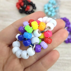 Pode incluir: Um brinquedo fidget de contas colorido e feito à mão. O brinquedo apresenta uma variedade de contas brilhantes coloridas do arco-íris enfiadas em anéis de metal. Outros brinquedos de contas em preto, azul e roxo são visíveis no fundo.