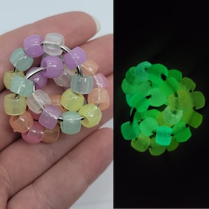 Pode incluir: Um anel de contas com contas translúcidas de cores pastel enfiadas em um anel de metal prateado. As contas brilham em verde brilhante no escuro. O anel é mostrado em duas condições de iluminação diferentes.
