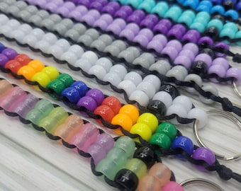 May include: A close-up of a rainbow of colorful beaded keychains. Each keychain is made of small, round beads strung on a black cord. The keychains are arranged in a rainbow pattern, with the colors fading from red to purple. The keychains are attached to silver key rings.