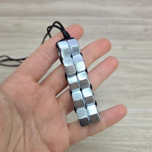 May include: A handmade necklace featuring a series of silver metal nuts strung together with black cord. The necklace is held in a hand against a light wood grain background.