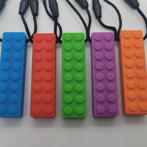 Puede incluir: Cinco juguetes masticables de silicona con forma de ladrillo Lego, cada uno en un color diferente: azul, naranja, verde, morado y naranja. Cada juguete tiene una cuerda negra atada.