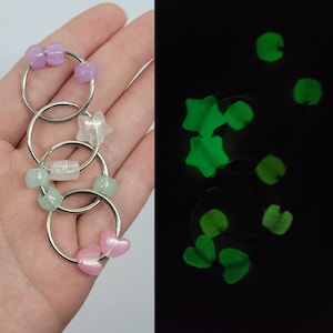 Pode incluir: Um conjunto de quatro chaveiros prateados, cada um adornado com contas e berloques coloridos. Os berloques incluem estrelas, corações e formas retangulares. Os chaveiros brilham em verde no escuro.