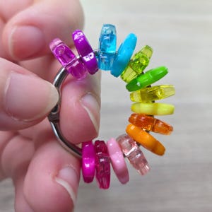 May include: A silver-toned metal ring holding a collection of small, translucent, and opaque plastic beads in a spectrum of rainbow colors including purple, blue, green, yellow, orange, and pink, arranged in a circular pattern on the ring.