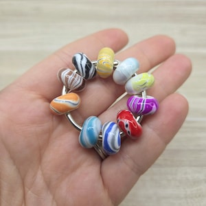 May include: A collection of colorful, patterned beads strung on a silver ring. The beads feature a variety of colors including orange, blue, yellow, red, and purple, with swirled and striped designs. The beads are held in a hand.