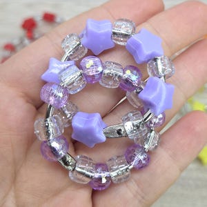 May include: A bracelet with a combination of purple star-shaped beads and clear, iridescent beads. The bracelet is held together by a silver-coloured metal ring. The beads are arranged in a circular pattern, creating a playful and colourful accessory.