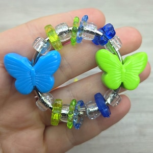 May include: A circular ring featuring two large plastic butterfly charms in bright blue and lime green. The ring is decorated with a variety of smaller beads, including clear faceted beads, translucent blue and green flower-shaped beads, and small lime green rings.