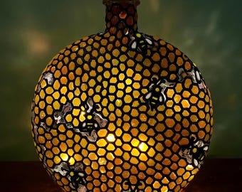 May include: A decorative glass bottle with a honeycomb pattern in yellow and black. The bottle features a cork stopper and is illuminated from within, creating a warm glow. Several bee designs are incorporated into the mosaic pattern.