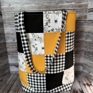 May include: A patchwork tote bag with a bee theme. The bag features a mix of black and white gingham, black, yellow, and floral patterned fabric. The bag has a black and white gingham strap.