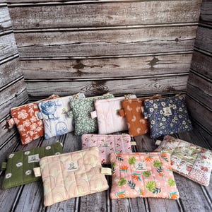 May include: A collection of quilted pouches in various colors and patterns, including floral, geometric, and abstract designs. Each pouch features a small fabric loop and a tag with the text "Cozy Creations". The pouches are arranged on a wooden surface.