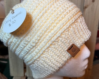 Op de afbeelding: Crèmekleurige, handgebreide beanie-muts. De muts wordt getoond op een mannequinhoofd. De muts heeft een geribbeld ontwerp en een klein leren label. Op het label staat "Cozy Beanie". Het woord "Cream" staat in witte letters onder het mannequinhoofd.