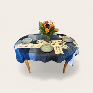 May include: An oval dining table with a patchwork denim tablecloth. The table is set with three gray plates, silverware, and a vase of orange flowers. The table has light brown, curved legs.