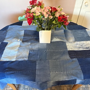 May include: A round denim patchwork tablecloth in various shades of blue covers a wooden table. A white square vase holds a bouquet of red and pink flowers. The table is set against a light pink wall.
