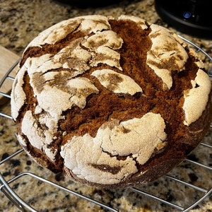 May include: A round loaf of freshly baked bread with a dark brown crust and a light tan, cracked top. The bread sits on a silver wire cooling rack, with a speckled countertop in the background.