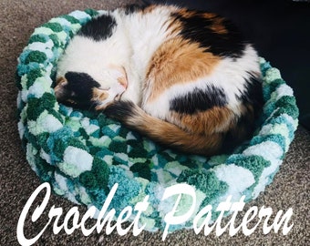 Puede incluir: Una cama para gatos hecha a crochet en tonos verdes y azules con un gato blanco y marrón durmiendo dentro. La cama está hecha con un hilo texturizado y tiene una forma redonda. Crochet Pattern.