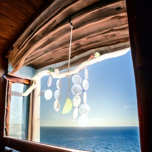 May include: A wooden branch with a hanging wind chime made of silver-colored metal discs. The wind chime is hanging in front of a window with a view of the ocean.