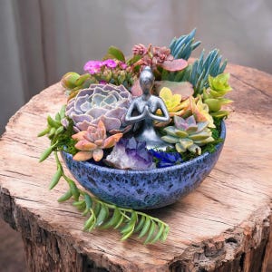 May include: A blue ceramic planter with a variety of succulents and a silver figurine of a person in a yoga pose. The planter is sitting on a wooden stump.