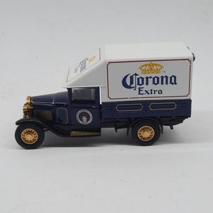 May include: A miniature dark blue vintage delivery truck featuring a white cargo box with the Corona Extra logo in blue and gold. The truck has gold-colored wheels, a gold front grille, and a circular logo on the door that reads Cerveceria Modelo.