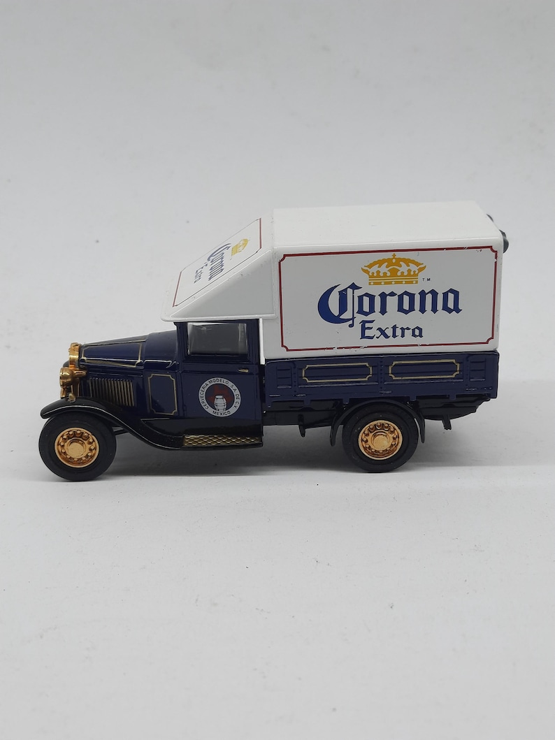 May include: A miniature dark blue vintage delivery truck featuring a white cargo box with the Corona Extra logo in blue and gold. The truck has gold-colored wheels, a gold front grille, and a circular logo on the door that reads Cerveceria Modelo.