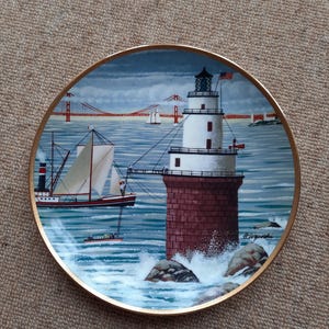 Op de afbeelding: Decoratieve plaat met een vuurtorenscène met een rode bakstenen basis en witte toren, een zeilboot en de Golden Gate Bridge op de achtergrond. De plaat heeft een gouden rand en is geplaatst tegen een blauwe lucht en oceaan.