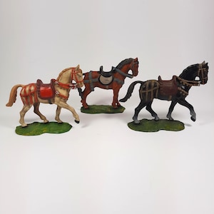 May include: Three toy horses with saddles and bridles. The horses are brown, white, and black. They are standing on green bases.