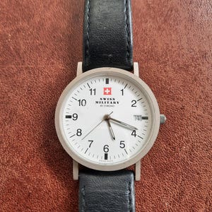 May include: A silver watch with a white face and black leather band. The watch face has black numbers and hands, a red Swiss cross, and the text "SWISS MILITARY BY CHRONO".