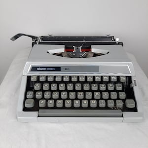 May include: A white Silver-Reed 100 typewriter with a black keyboard and a carriage return lever. The typewriter has a silver nameplate with the brand name and model number.