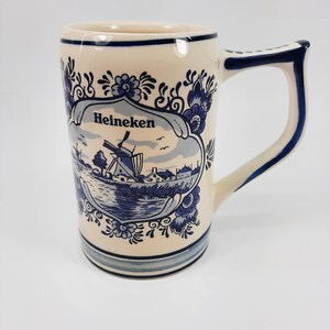 May include: Cream ceramic mug with a blue and white Dutch windmill design. The mug features the word "Heineken" and floral accents. The handle and rim are trimmed in blue. A decorative piece for beverages.