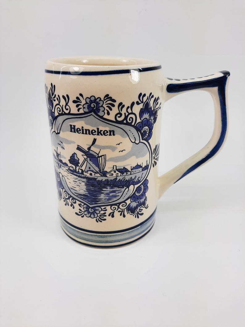 May include: Cream ceramic mug with a blue and white Dutch windmill design. The mug features the word "Heineken" and floral accents. The handle and rim are trimmed in blue. A decorative piece for beverages.