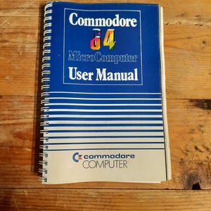 Può includere: Un manuale utente del Commodore 64 Microcomputer di colore blu con una copertina a strisce bianche e blu e il logo Commodore Computer.