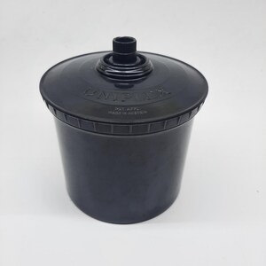Peut inclure: Une cuve de développement de film cylindrique noire avec un couvercle. Le couvercle présente un bouton central et le texte en relief UNIPLEX, PAT APPL et MADE IN AUSTRIA. Le récipient a une finition mate lisse et une forme légèrement conique.