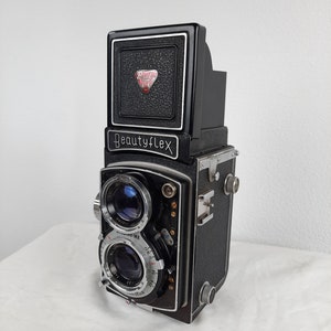 May include: A black Beautyflex twin-lens reflex camera with silver accents. The camera has two lenses, a top viewfinder, and a textured body. The name "Beautyflex" is printed on the front. The camera is on a white surface.