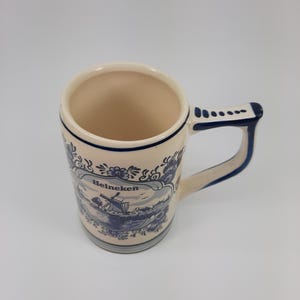 May include: Cream ceramic mug with a blue and white landscape design and the word "Heineken." The mug has a rounded body, a sturdy handle, and a dark blue trim. The design features a windmill and water scene.