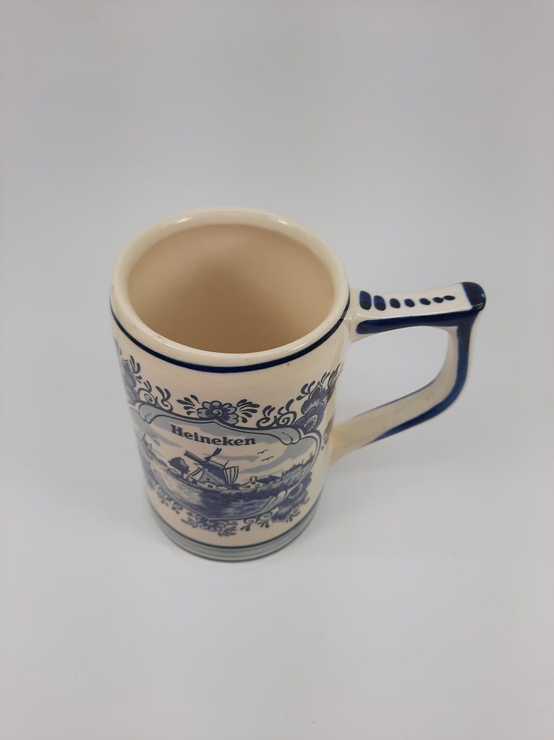 May include: Cream ceramic mug with a blue and white landscape design and the word "Heineken." The mug has a rounded body, a sturdy handle, and a dark blue trim. The design features a windmill and water scene.
